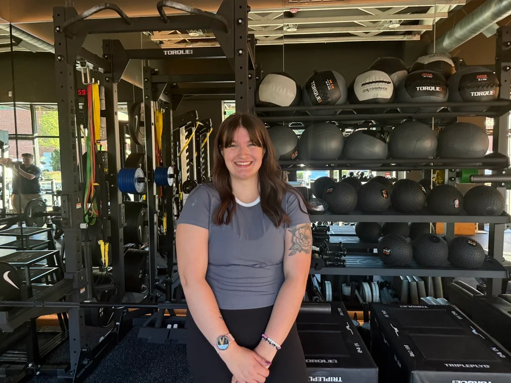 Portland personal trainer Jordyn Tietz smiling in a gray T-shirt and black leggings, standing in front of a rack filled with medicine balls inside a modern gym.