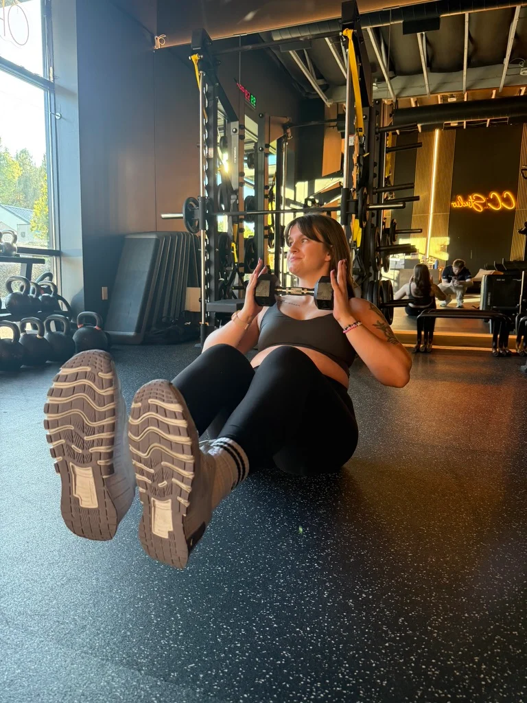 Portland personal trainer Jordyn Tietz in a black sports bra and leggings performing a seated ab crunch on the gym floor, feet lifted and hands raised, with the CC Studio neon sign glowing in the background.