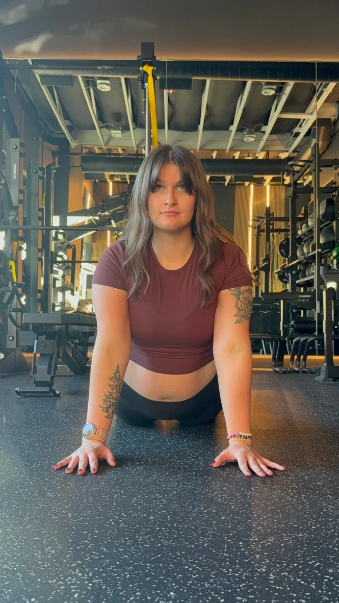 Portland personal trainer Jordyn Tietz in a maroon crop top holding a push-up plank position on a gym floor, looking directly at the camera with weight racks and training equipment behind her.
