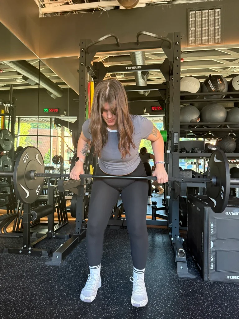 Portland personal trainer Jordyn Tietz in a gray T-shirt and black leggings gripping a loaded barbell at the start of a deadlift, with medicine balls and strength equipment visible behind her.