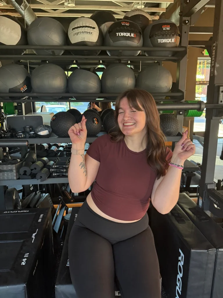Portland personal trainer Jordyn Tietz laughing with eyes closed in a maroon crop top, both hands raised with fingers crossed, in front of a rack of medicine balls in a bright, open gym.