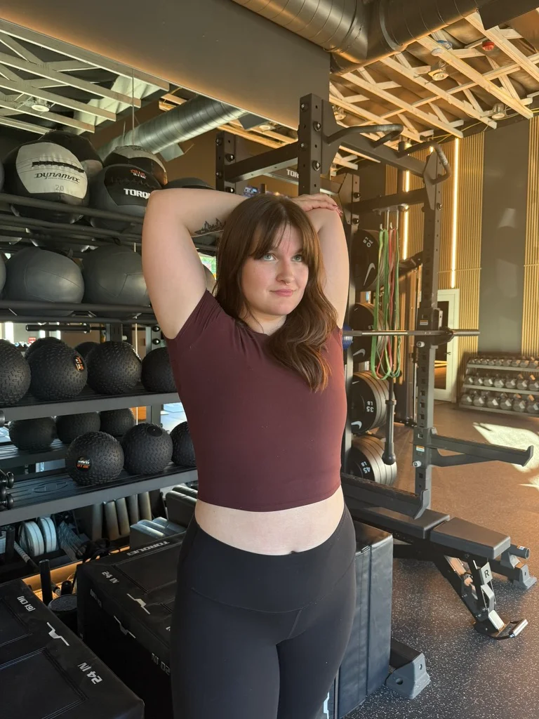 Portland personal trainer Jordyn Tietz in a maroon crop top and black leggings performing an overhead arm stretch in a gym, with racks of medicine balls and resistance bands behind her.