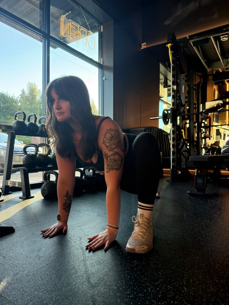 Portland personal trainer Jordyn Tietz in a black sports bra and leggings in a bear crawl position on a gym floor, hands planted and gaze forward, with kettlebells and large windows letting in natural light behind her.