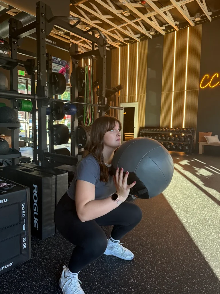 Portland personal trainer Jordyn Tietz in a gray T-shirt and black leggings performing a deep squat while holding a medicine ball at her chest, with gym equipment and the CC Studio neon sign glowing in the background.
