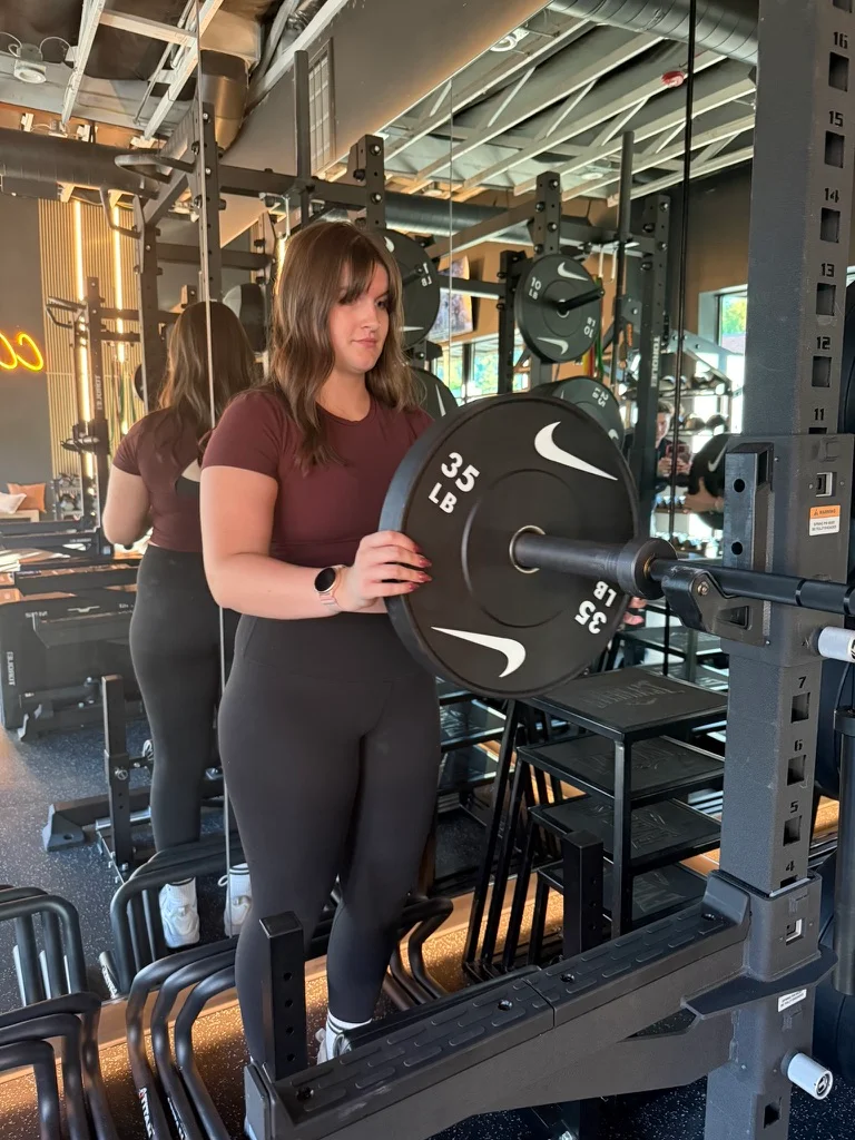 Portland personal trainer Jordyn Tietz in a maroon T-shirt and black leggings loading a 35-pound weight plate onto a barbell in a squat rack, with her reflection visible in the gym's floor-to-ceiling mirrors.